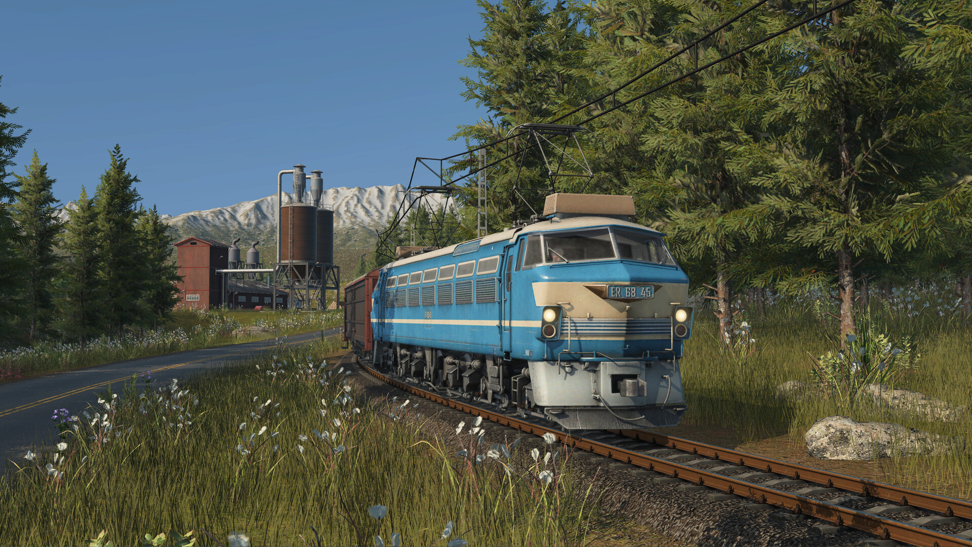 A blue electric locomotive hauling freight wagons past an industrial silo in Transport Fever 3