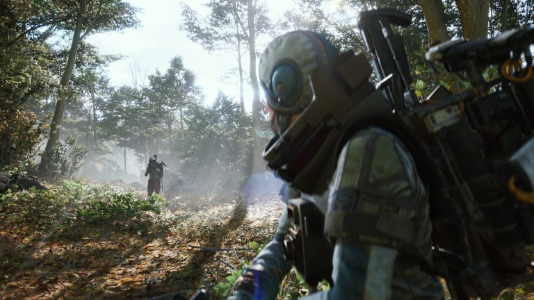Over the shoulder of a helmeted raider in a sunlit forest, looking at another raider with a large backpack walking toward them along a leafy path
