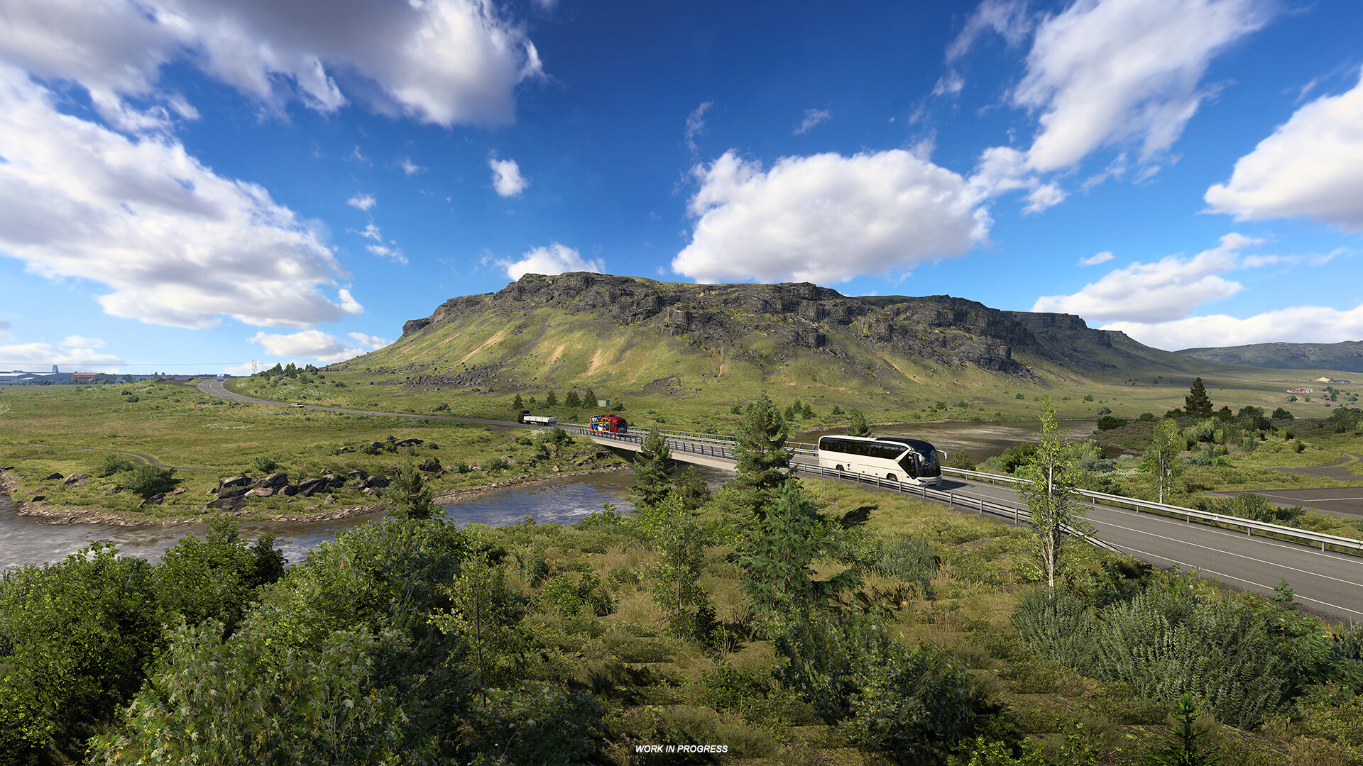 A coach crossing a river bridge below a flat-topped hill under a blue, cloudy sky in Euro Truck Simulator 2: Iceland, marked Work in Progress.
