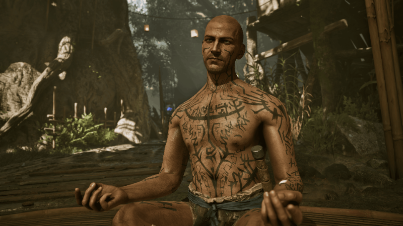 A bald man covered in dark tattoos sits cross-legged among candles in the Swamp Camp