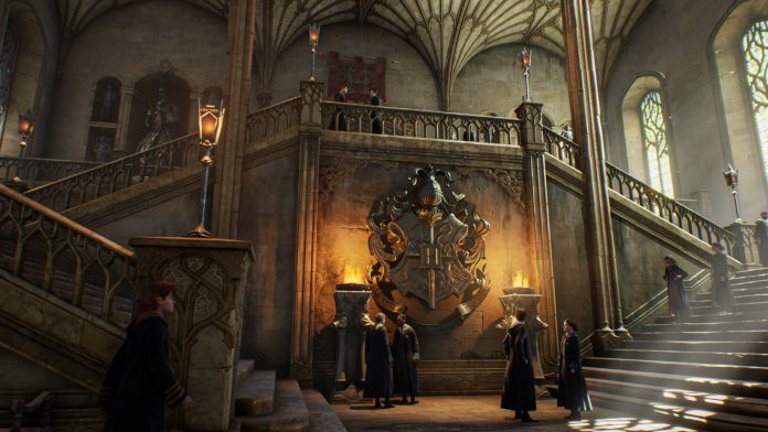 Students in dark robes standing in a stone entrance hall in Hogwarts Legacy, beneath curving staircases and a large carved crest lit by two braziers.