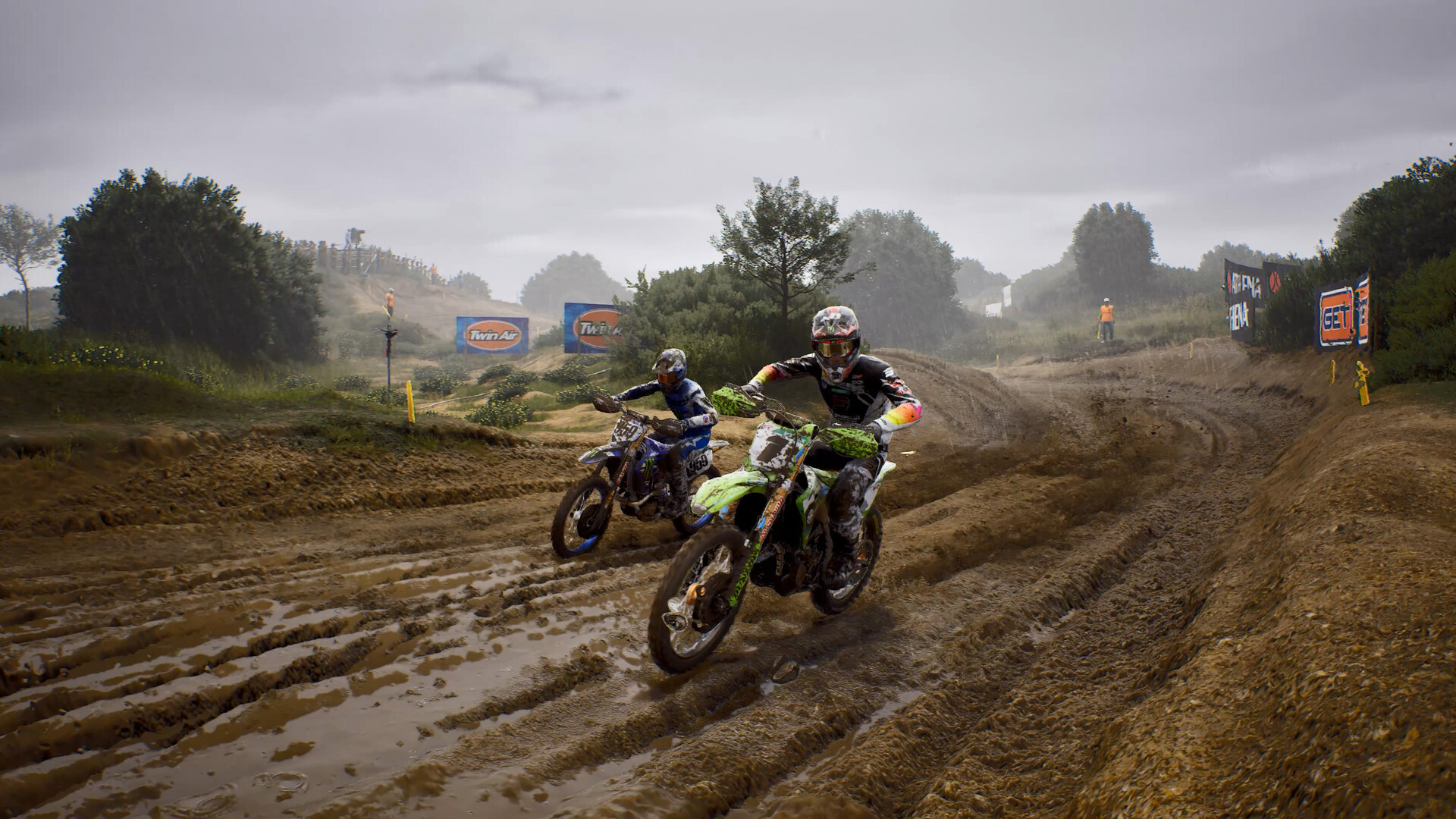 Two motocross riders race along a muddy, rutted track lined with trees