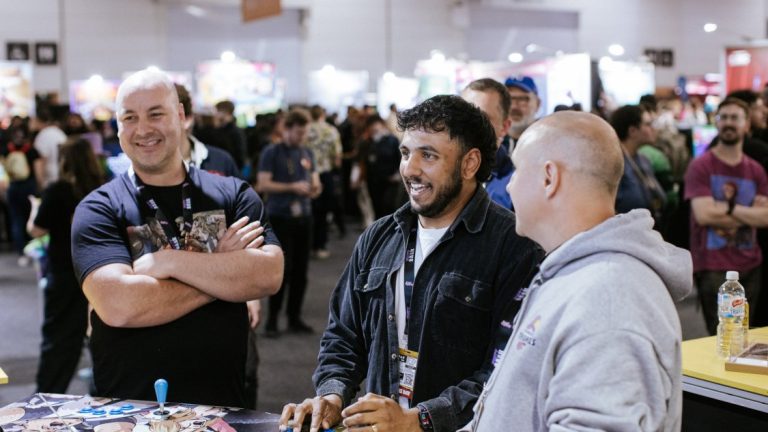 Three smiling men chat at a booth table in the busy PAX Aus expo hall