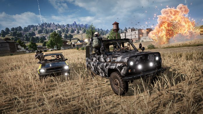 Two armed players ride in a camouflaged jeep and a sedan across a grassy field as an explosion goes off near a water tower