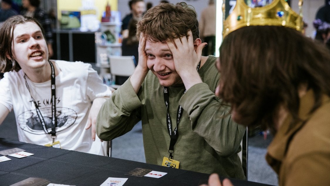 Two young players with lanyards react with mock horror at a table game while someone in a crown looks on