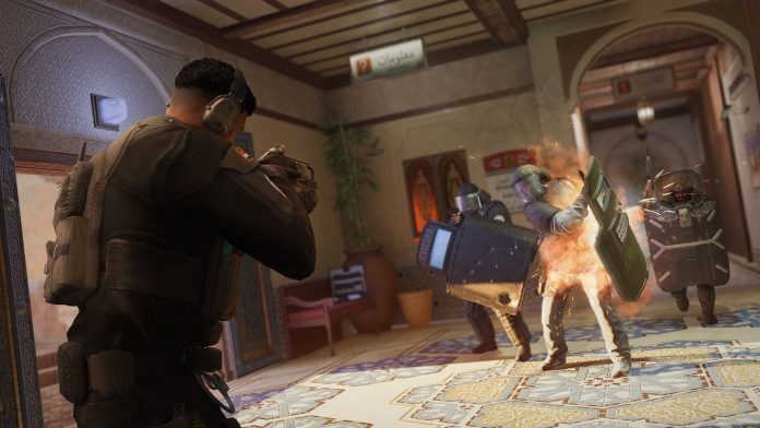 Rainbow Six Siege breach. Image: Ubisoft Attackers with ballistic shields push into a tiled hallway as an explosion bursts near a defender in Rainbow Six Siege