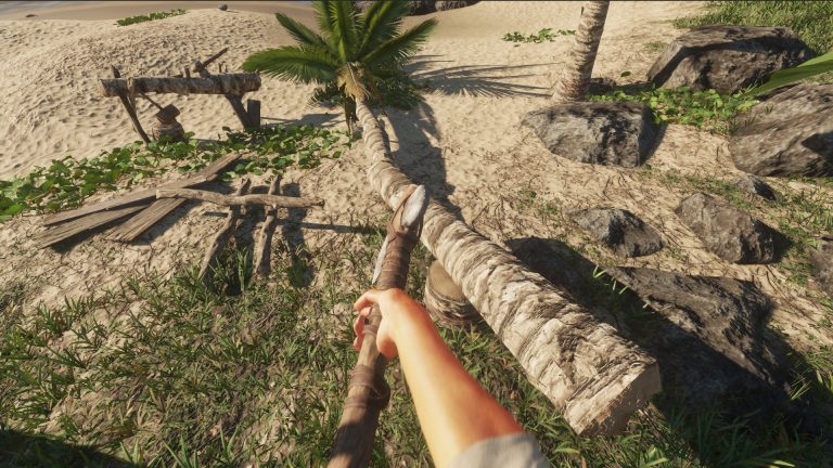 First-person view in Stranded Deep of a hand holding a spear on a sandy beach, beside a felled palm trunk and a wooden crafting frame.