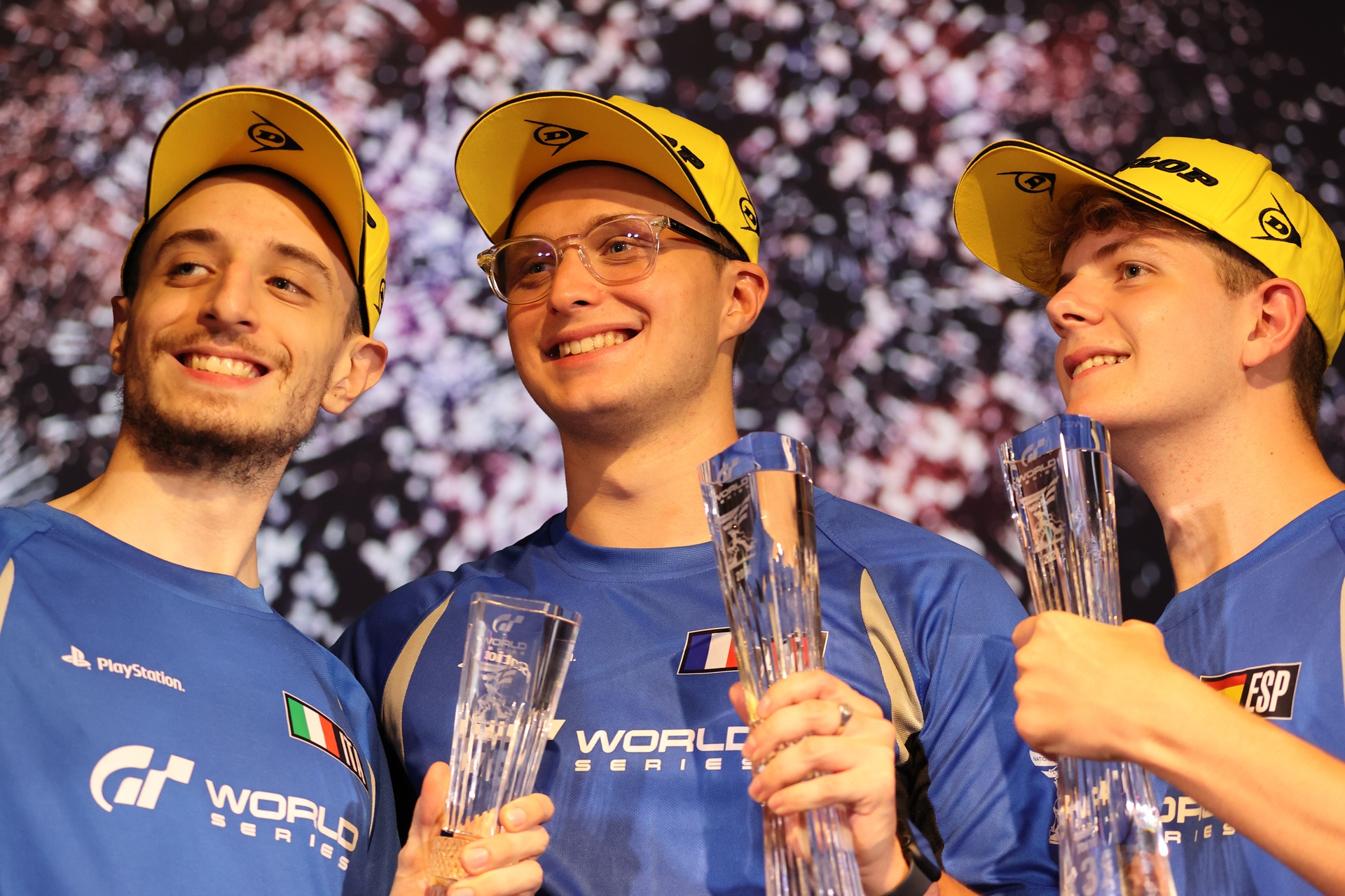 Three Gran Turismo World Series drivers in caps smile and hold trophies on a podium