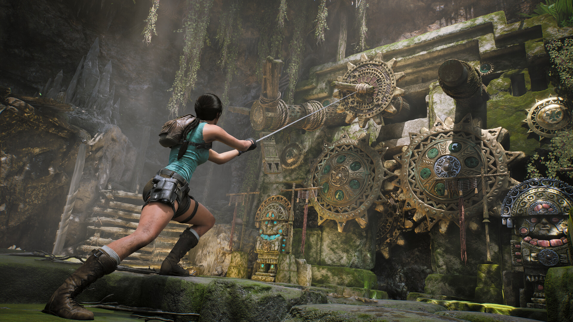 Lara Croft hauls on a rope attached to carved bronze gear wheels beside stone idols in an overgrown cave.